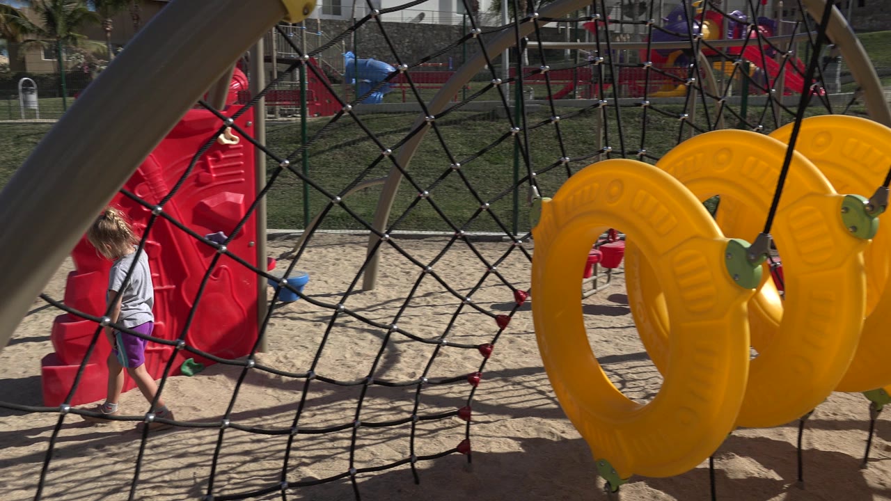 The child remains near the red playground equipment in San Jose del Cabo while the foreground rope net and yellow seats fill much of the frame.