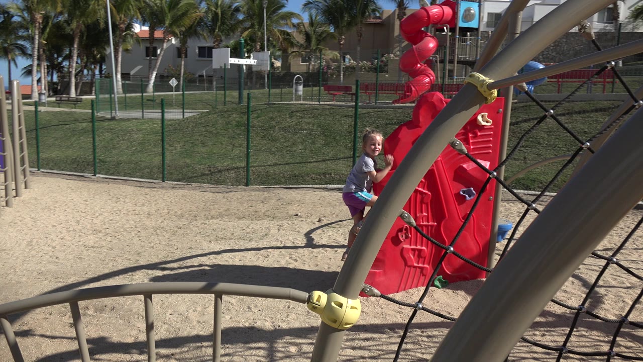 A little girl in a gray shirt and bright shorts climbs a red playground panel beside a black rope net in San Jose del Cabo, Mexico, with sand and a fenced park behind her.