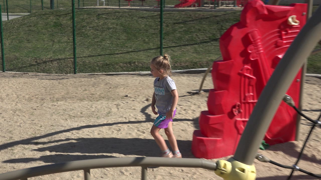 A young girl walks across a sandy playground in San Jose del Cabo, Mexico, with red play equipment and green fencing behind her.