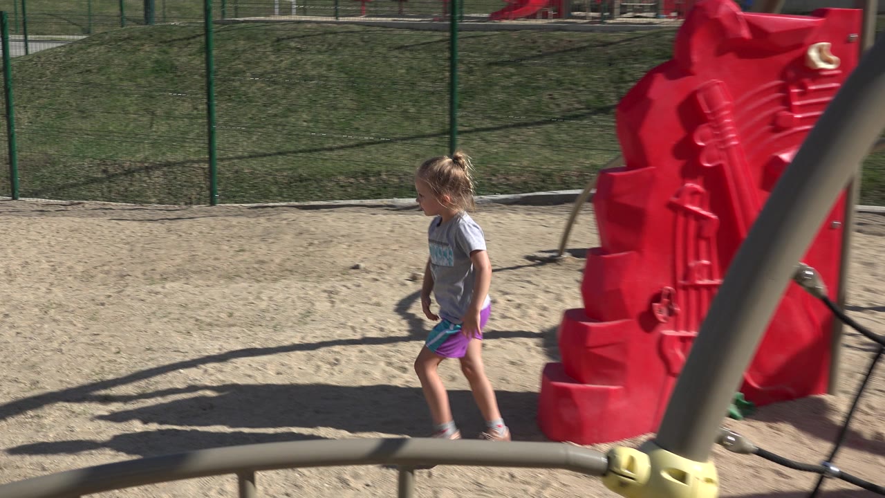 The girl continues walking on the sand near the same red playground structure, with a curved beige bar crossing the foreground in San Jose del Cabo, Mexico.