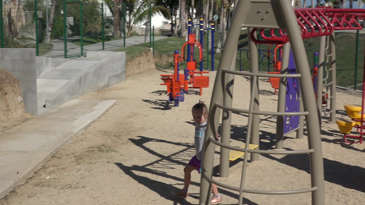Little girl partly behind a tan playground climbing frame on sandy ground.