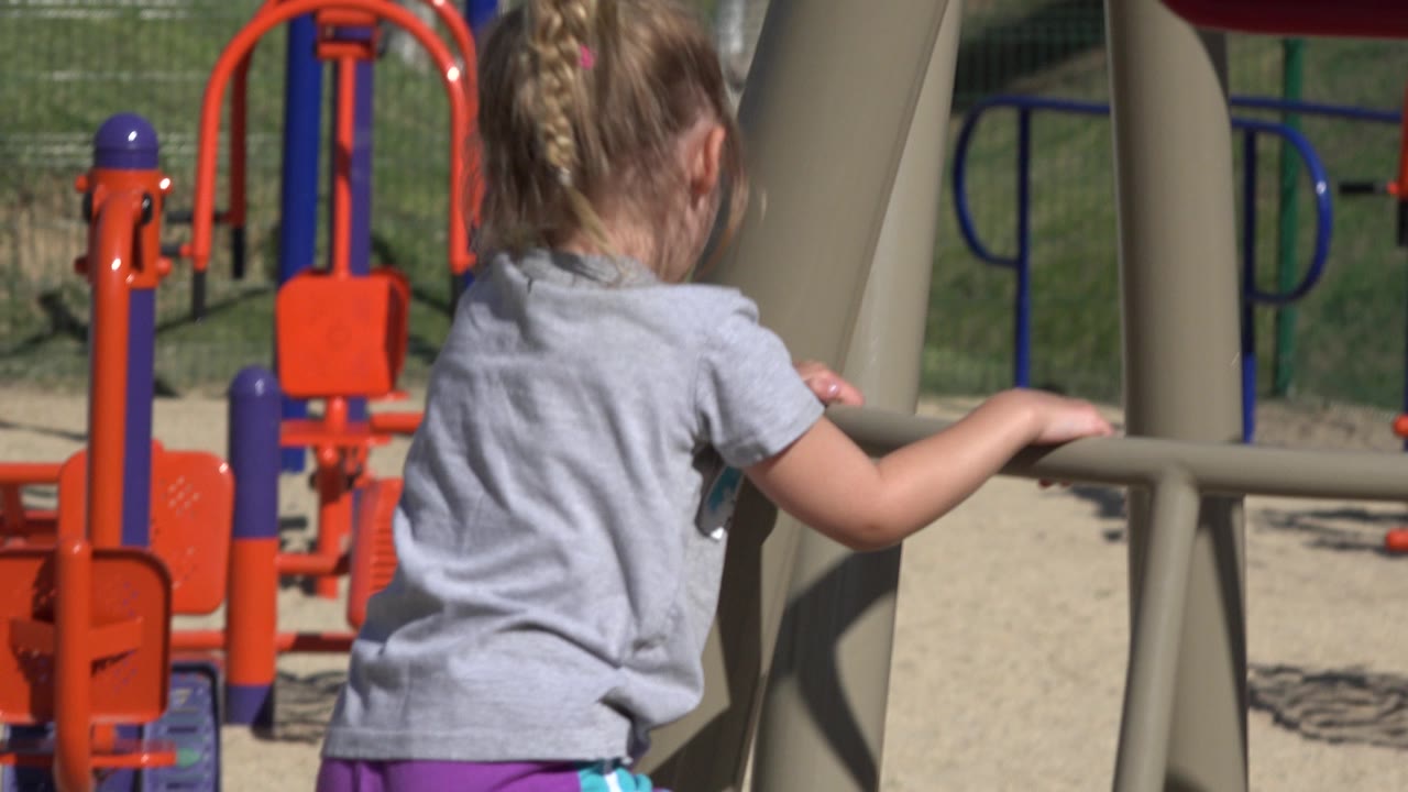 The child grips a beige playground support from behind in San Jose del Cabo, with red-orange play equipment and blue rails visible in the sunlit background.