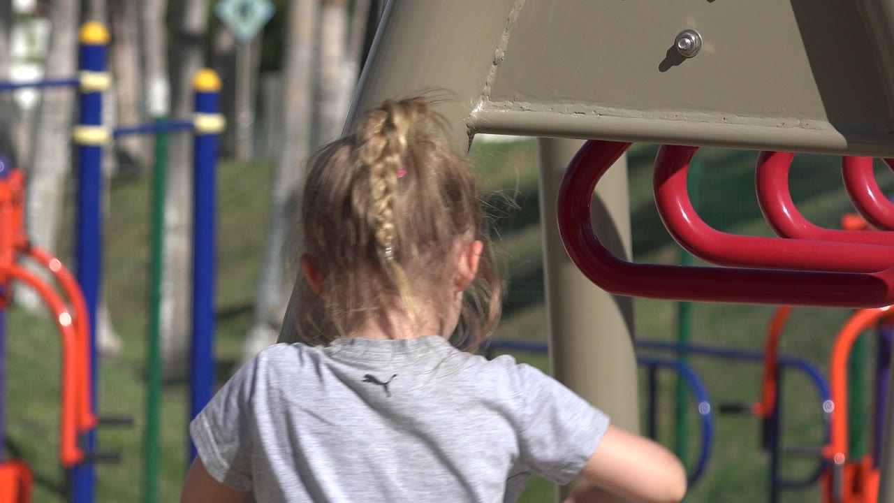 The child remains framed from behind beneath the beige play structure in San Jose del Cabo, with red bars crossing the upper right and soft-focus playground equipment in the background.