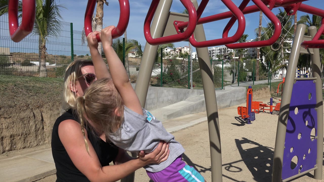 Woman supports a child hanging from red playground bars.