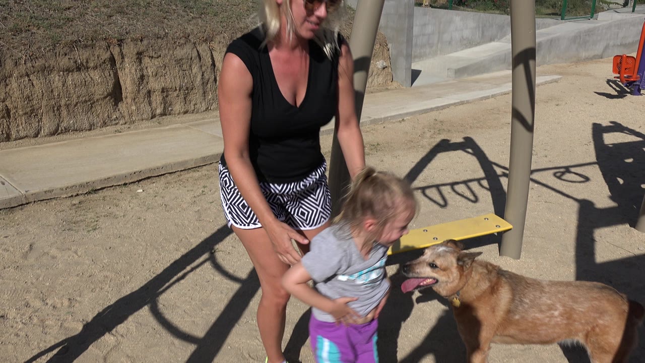In San Jose Del Cabo, Mexico, an adult woman stands beside a young girl and a brown dog near yellow and purple playground equipment in hard sunlight.