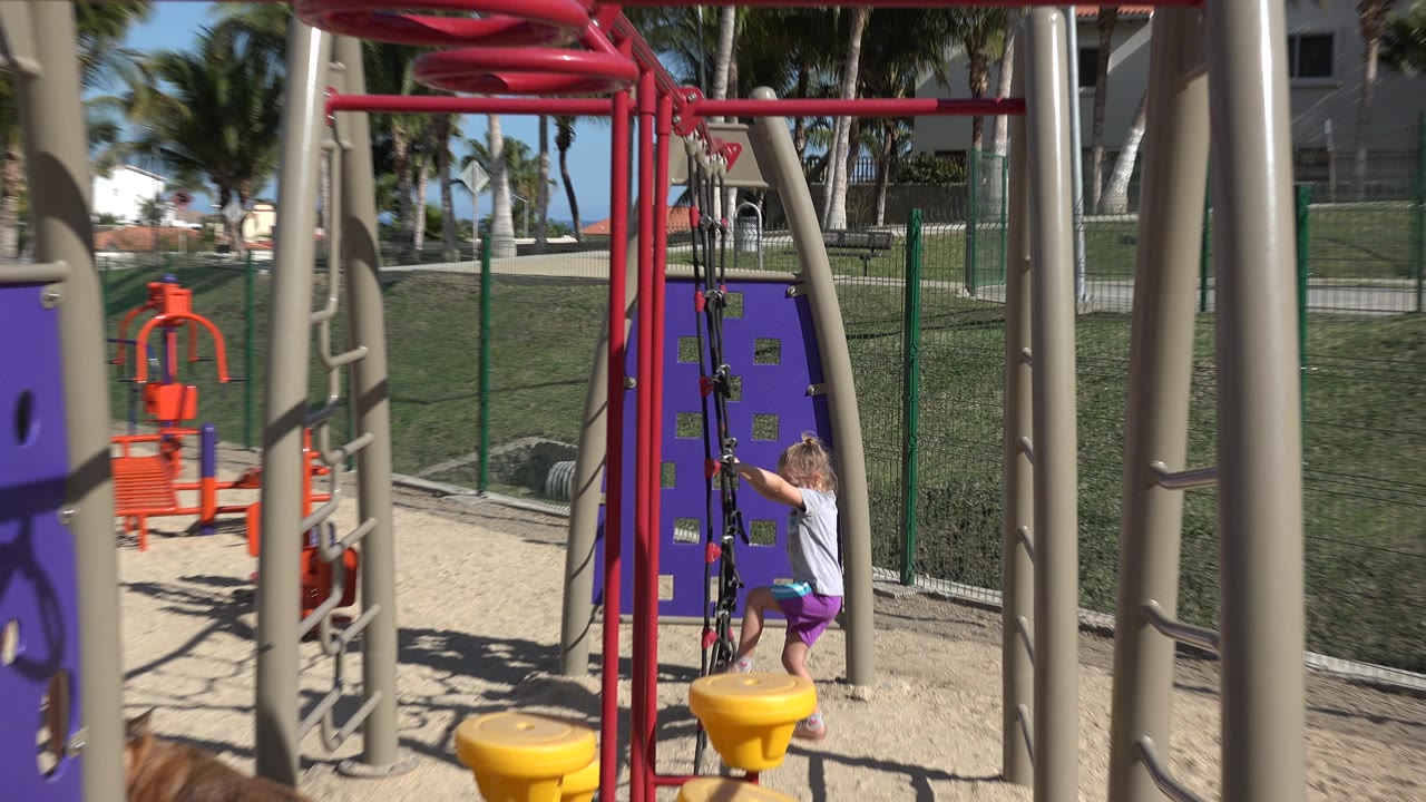 Wide playground frame with the girl higher on the rope net, red bars overhead, yellow play seats below, and fencing behind her in San Jose del Cabo, Mexico.