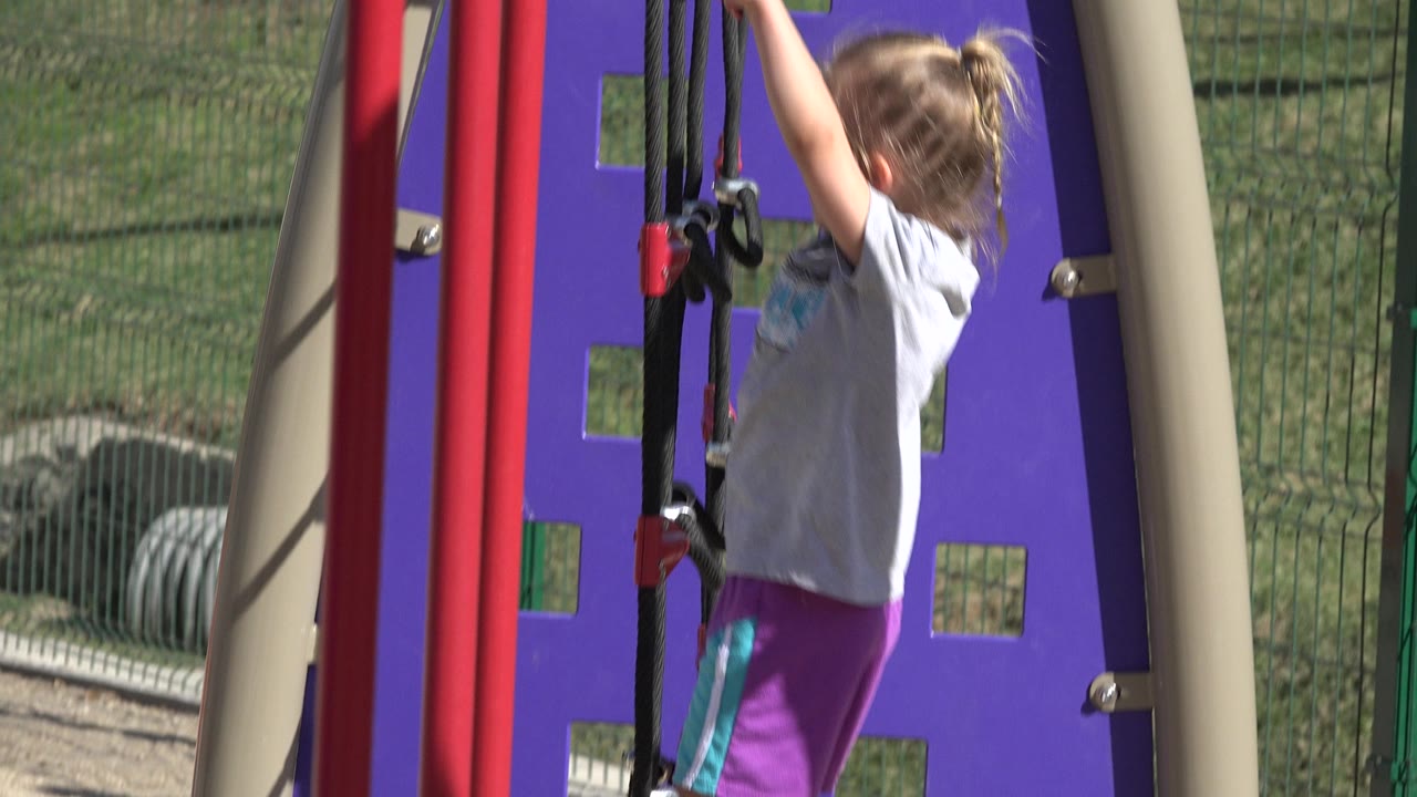 Young girl reaches up to playground ropes beside a purple panel.