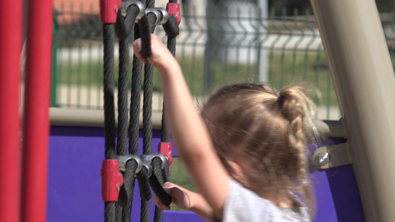 Blonde child reaches up on black playground ropes.