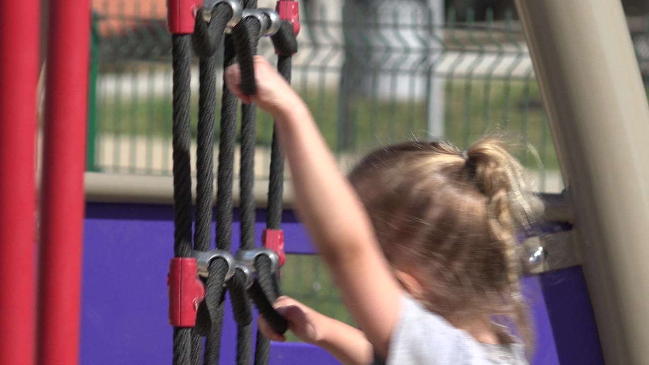 The child stays close to the black rope frame, one hand high and one hand lower, with red hardware and a fence behind the playground.