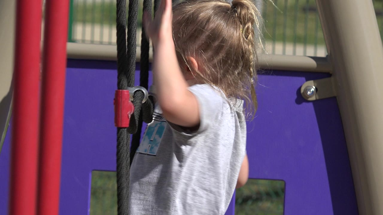 A young child in a gray shirt climbs black playground ropes next to red posts and a purple play panel in San Jose Del Cabo, Mexico.