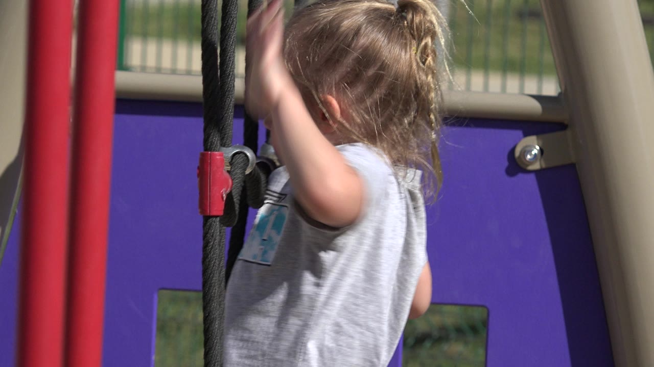 The child remains in close side view while holding the black ropes, with red posts and the purple playground wall behind her in San Jose Del Cabo, Mexico.