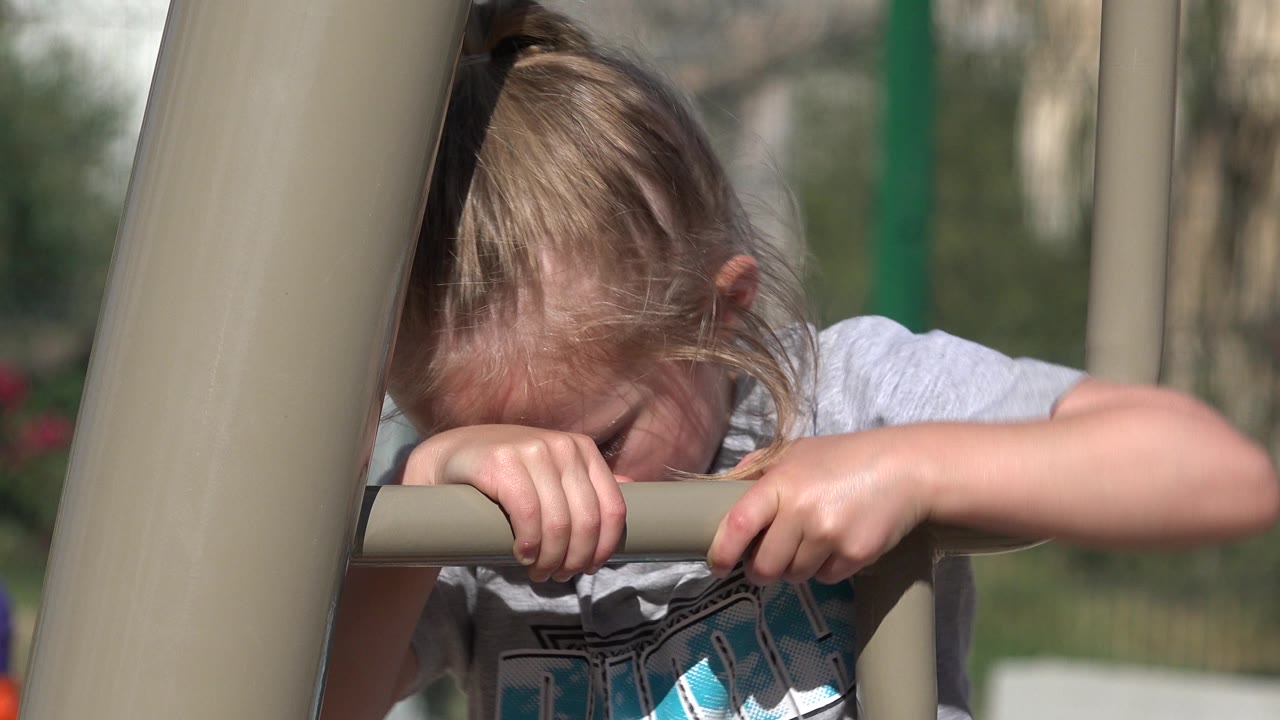 At 0:00, a young blond girl in San Jose del Cabo, Mexico grips a tan monkey bar with both hands while her face is partly hidden by the rail and a foreground post.