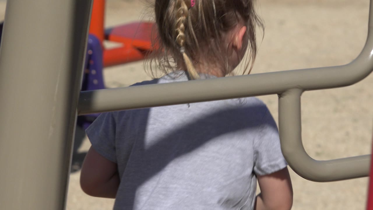 A young girl in a gray shirt is seen from behind at the San Jose del Cabo playground, partly obscured by rounded tan bars in strong sun.