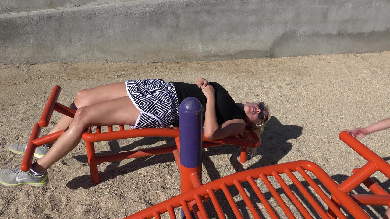 The woman remains reclined on the orange bench in San Jose del Cabo, Mexico, with one arm low and the orange exercise bars crossing the sandy foreground.