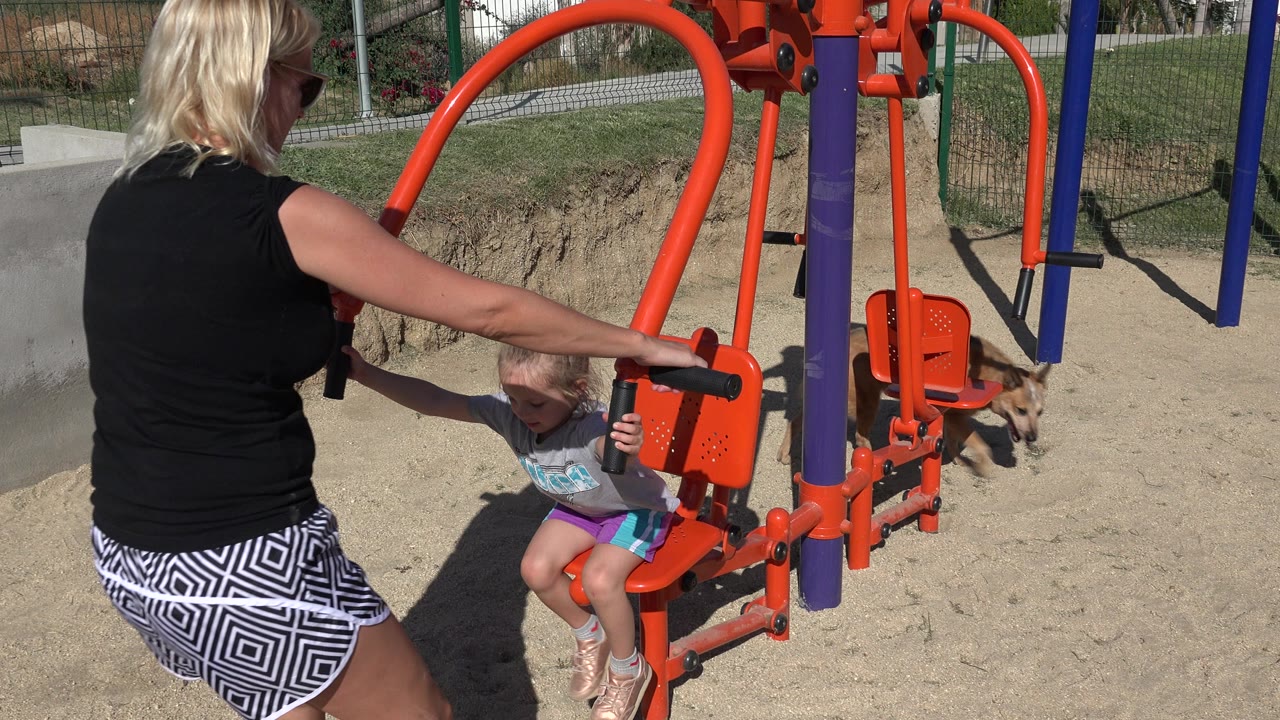 In San Jose del Cabo, Mexico, a blonde woman steadies a little girl seated on orange playground equipment while a tan dog walks in the sandy background.