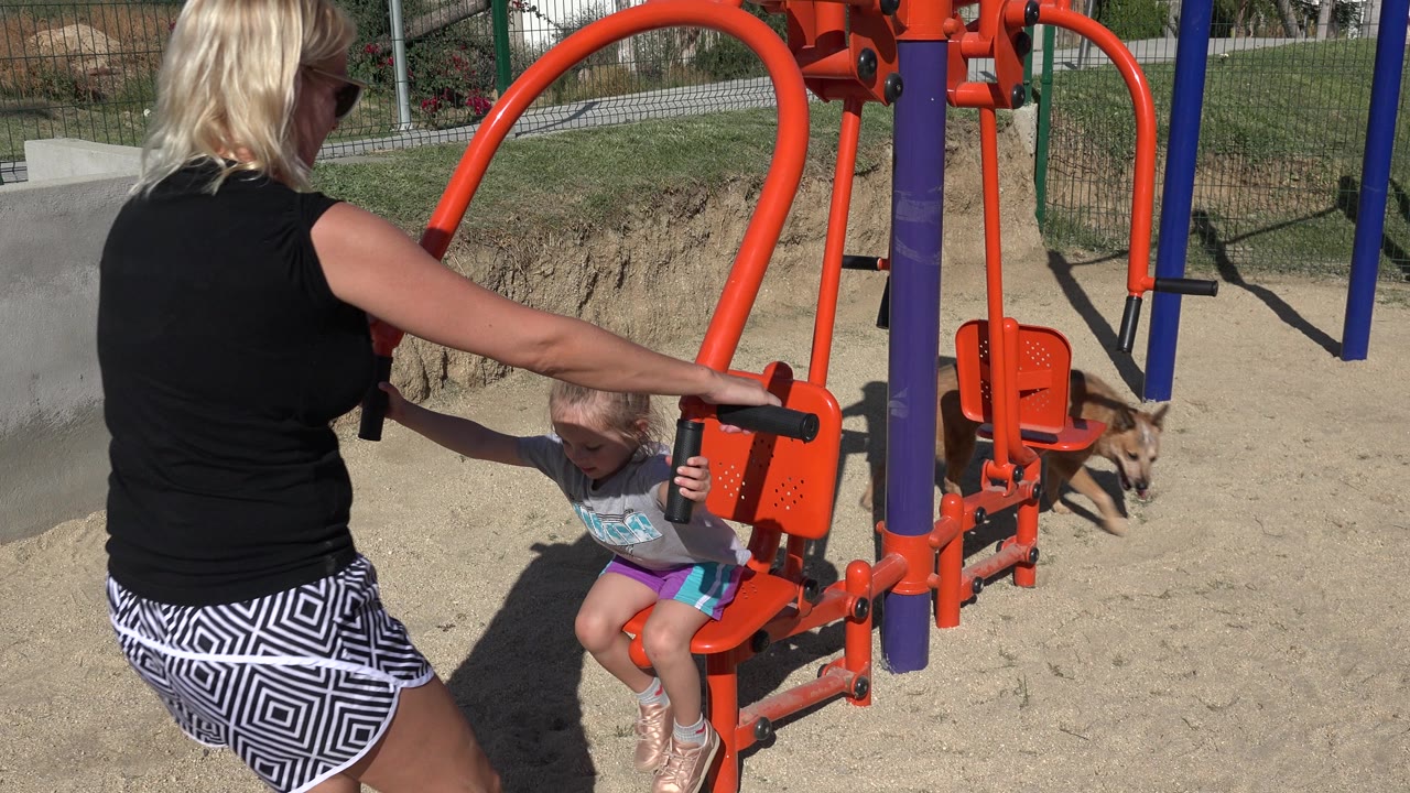 In San Jose del Cabo, Mexico, the woman reaches across the orange play structure as the little girl grips a handle and the dog moves closer in the sandy park.