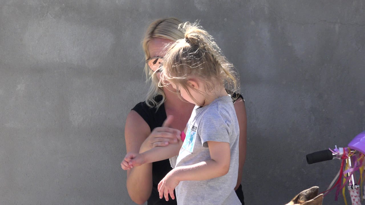 In San Jose del Cabo, Mexico, the woman looks down while helping the little girl with protective gear, with the gray wall filling the background and the scooter still partly visible.