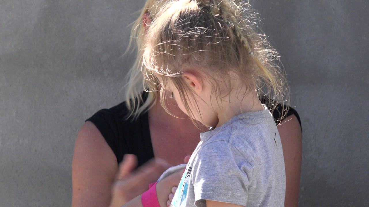 At 0:00 in San Jose Del Cabo, Mexico, a blonde child in a gray shirt stands close to a blonde woman in a black top against a gray wall.