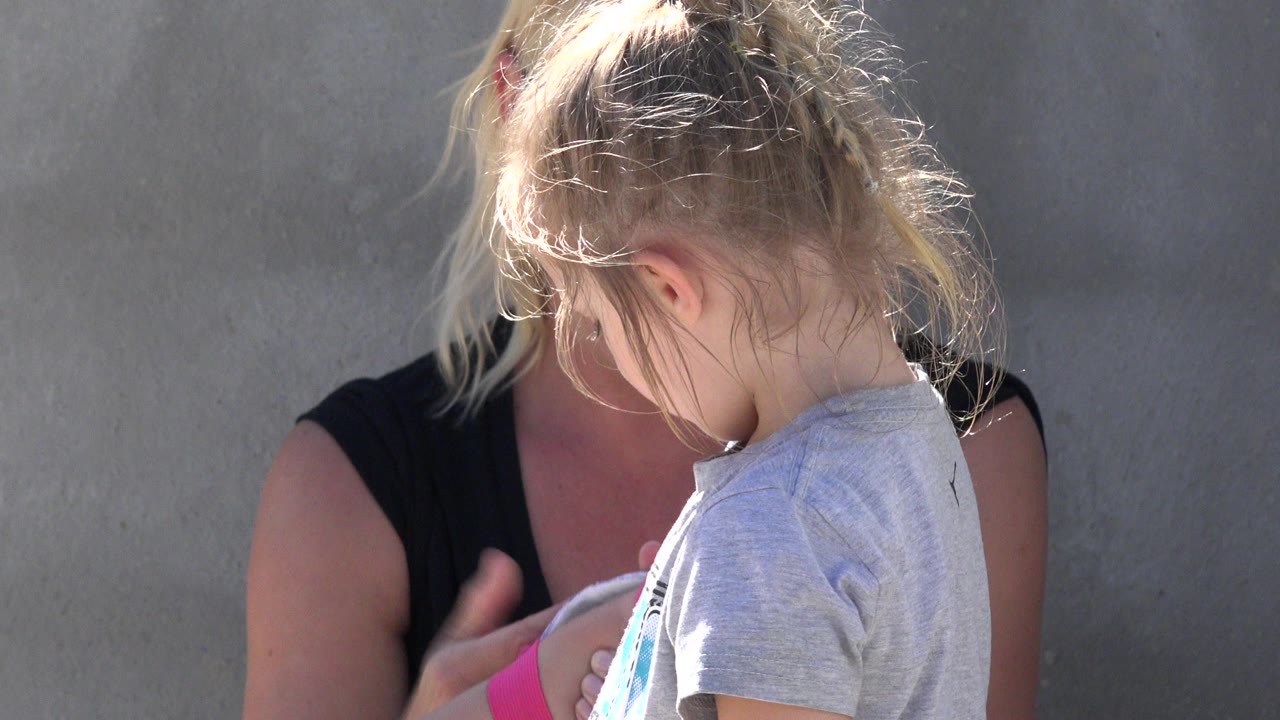 At 0:01, the child looks down in the foreground while the woman remains partly hidden behind her near the plain wall in San Jose Del Cabo, Mexico.