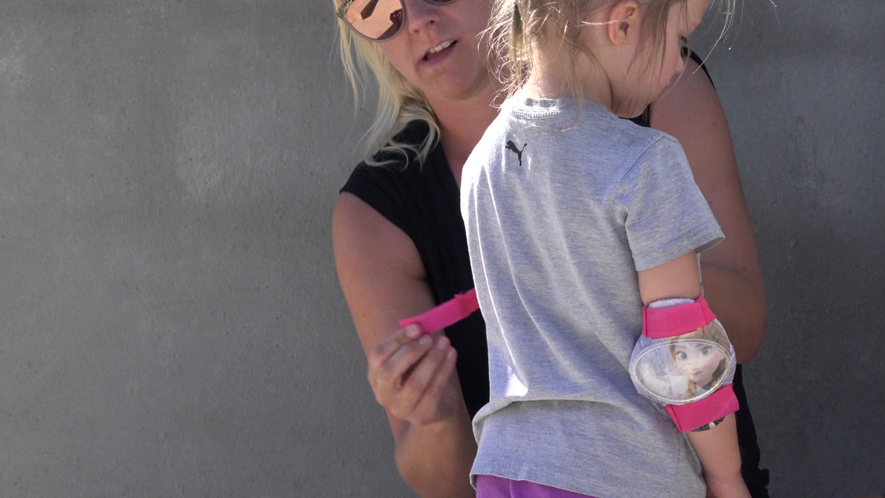 The mother remains partly behind the child, with the girl's back to camera and a pink elbow or wrist guard with cartoon artwork visible on her arm.