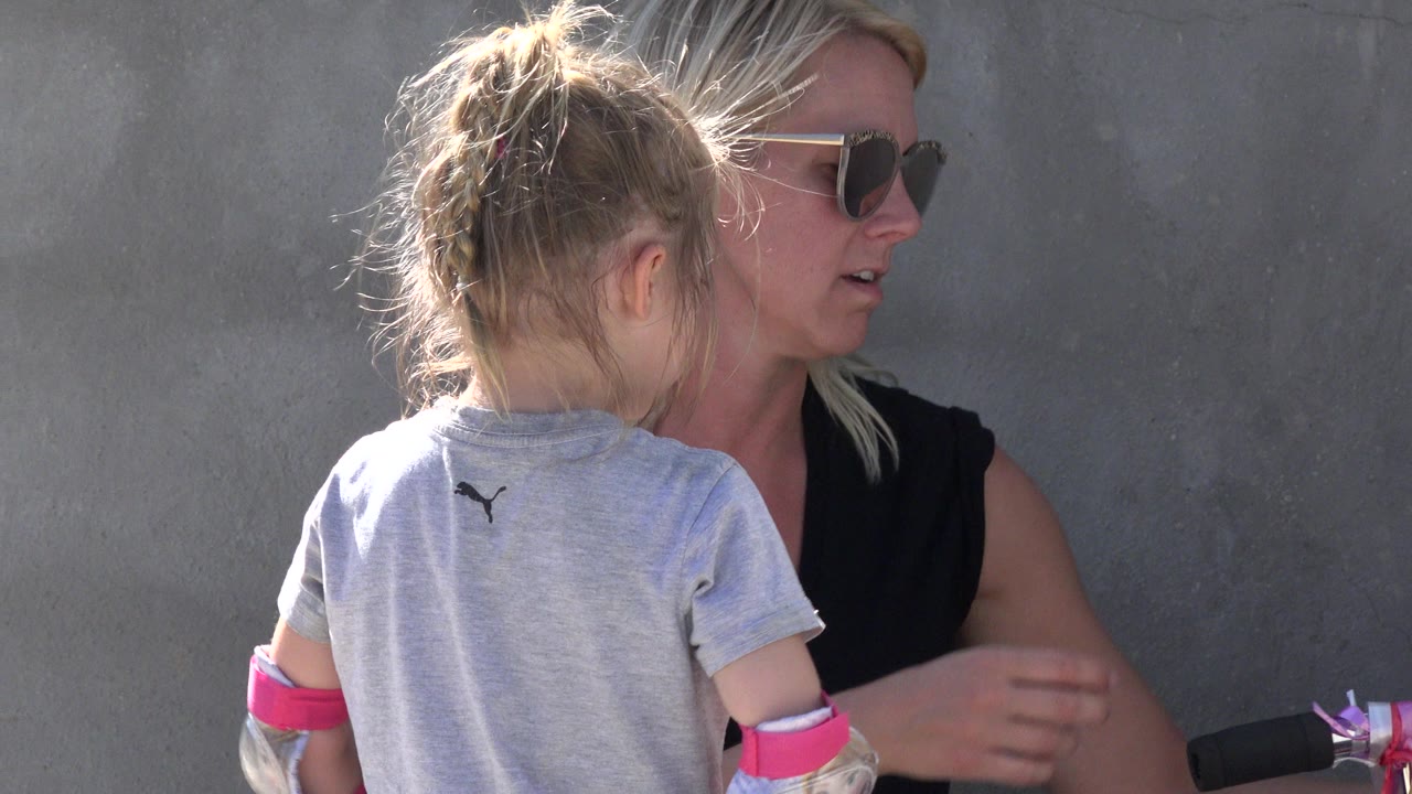 In San Jose del Cabo, a young girl faces away near an adult woman in sunglasses, with bright sun on their hair and a gray wall behind them.
