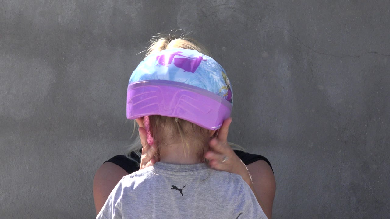 Child in gray shirt wearing a blue and purple helmet while an adult adjusts the straps.