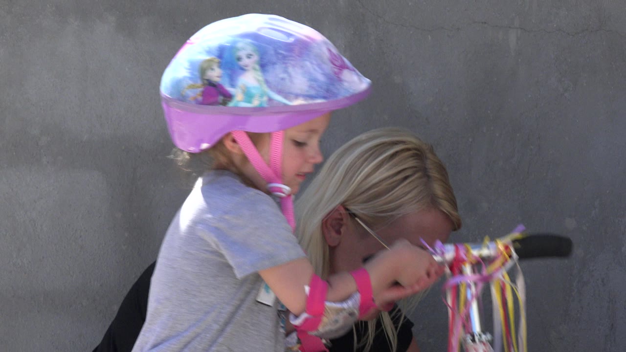 The girl in a purple helmet reaches toward the scooter's ribbon-covered handlebar while the adult woman remains bent close behind her in San Jose Del Cabo, Mexico.