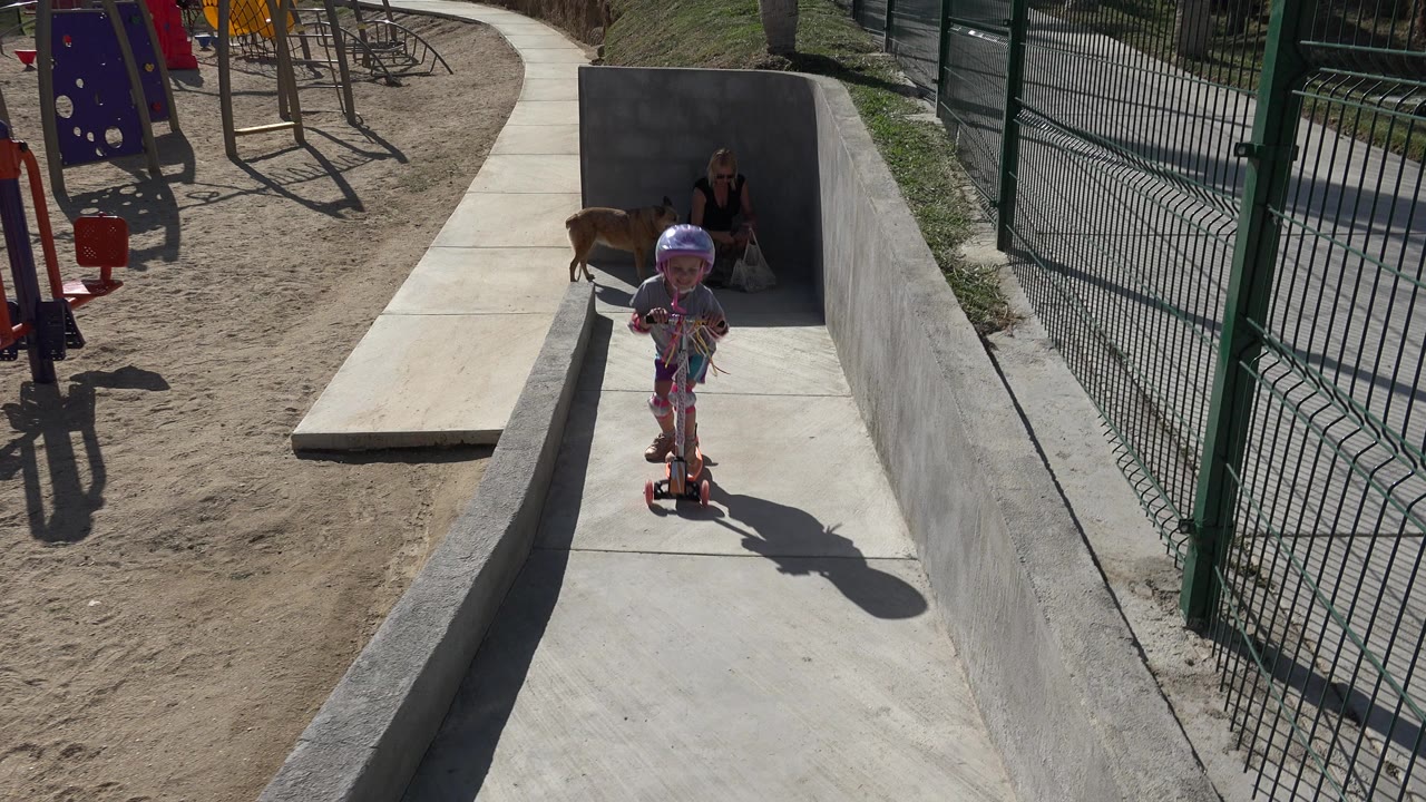 Child in a helmet rides a scooter down a concrete park path.