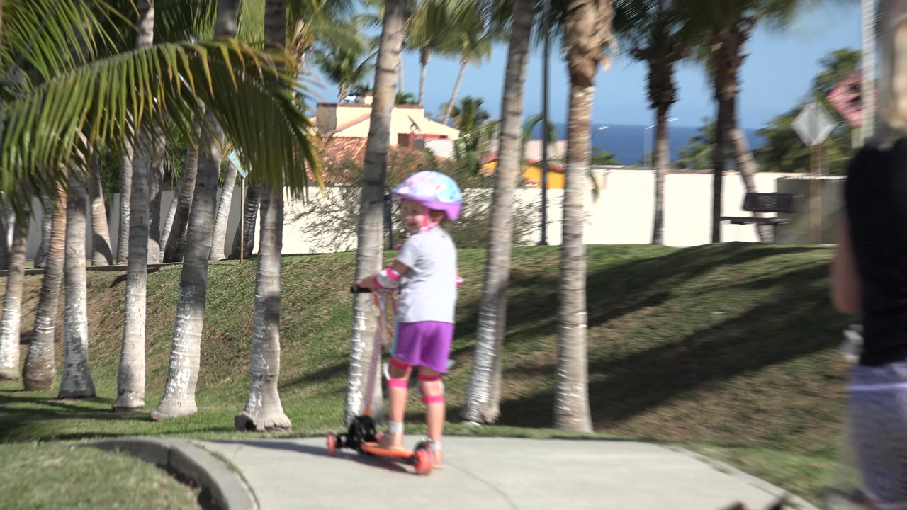 Young girl rides a scooter on a sidewalk lined with palm trees.