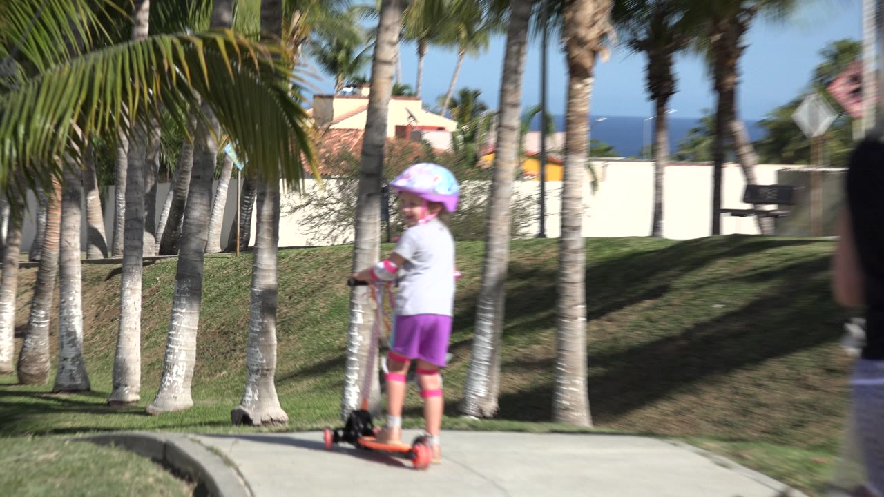The helmeted girl continues riding the scooter in San Jose del Cabo, Mexico, shown from behind and to the side as another child remains partially cropped at the right edge.