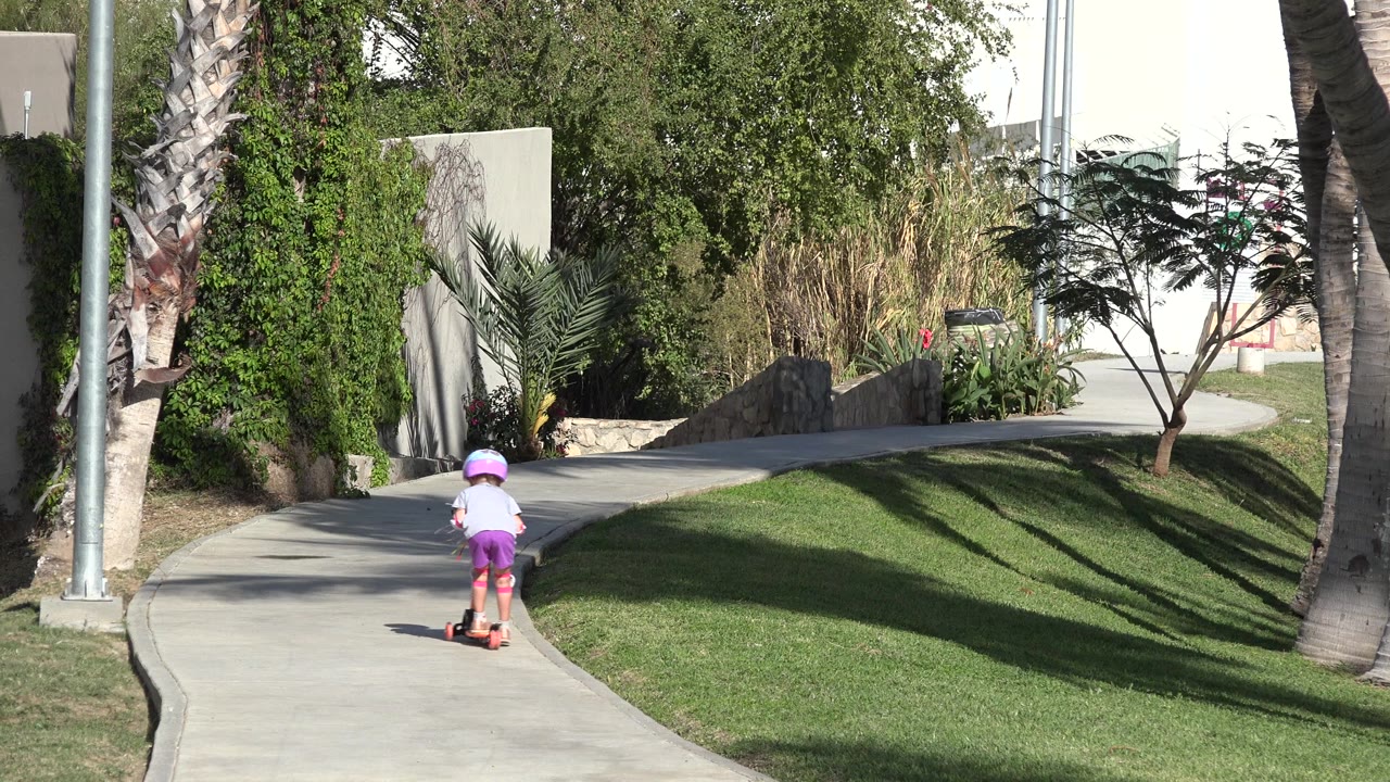 One second in, the child remains near the lower center of the San Jose del Cabo walkway, riding past a lawn edge with palms and planted shrubs ahead.