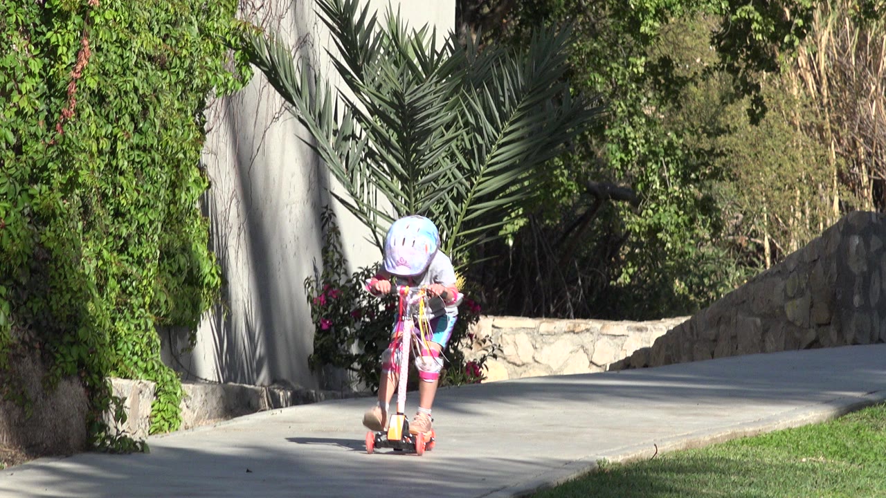 Young girl in a helmet rides a scooter on a sunlit path.
