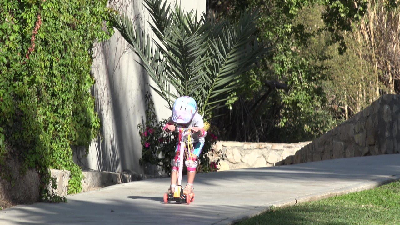 The child continues down the concrete path on a small scooter, framed by palms, dense green vines, and a low stone wall in San Jose del Cabo, Mexico.
