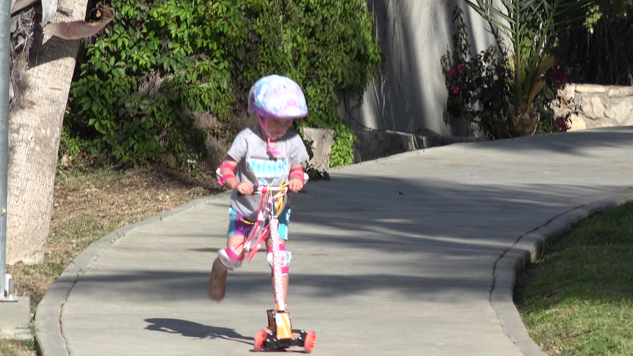 A child in a purple helmet and pads rides a decorated scooter toward the camera on a curved concrete path in San Jose del Cabo, Mexico.