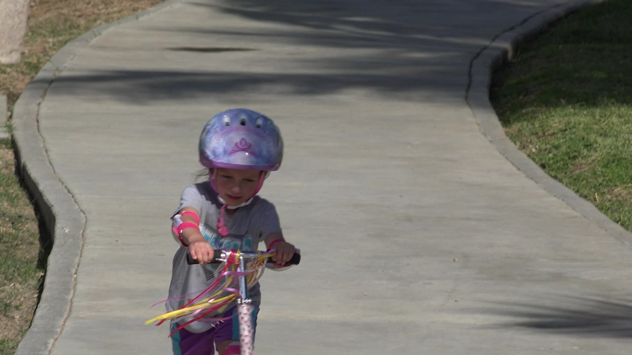 The girl stays centered as she rides closer on the concrete path, with grass and shadowed pavement behind her in San Jose del Cabo, Mexico.