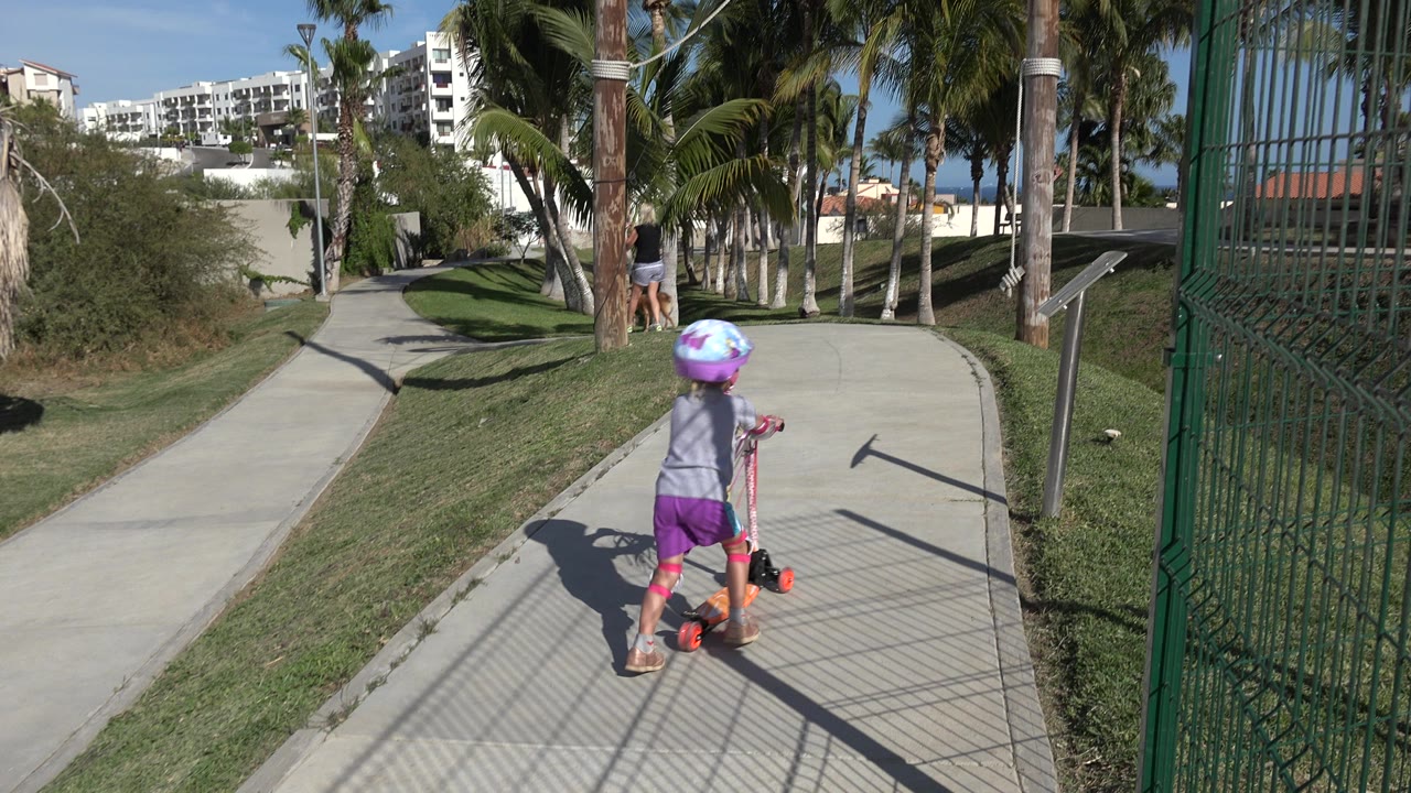 Young girl with purple helmet standing on an orange scooter on a sunny path.