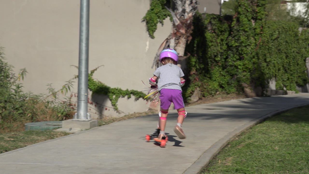 The child continues away from the camera on the orange three-wheeler, centered on the curving concrete path with grass to the right in San Jose del Cabo, Mexico.