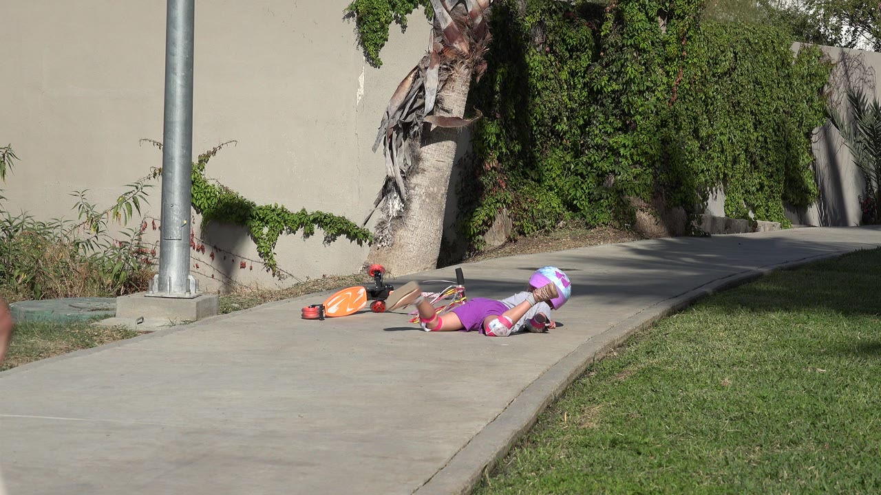 Helmeted child lying on a sidewalk beside an orange scooter.