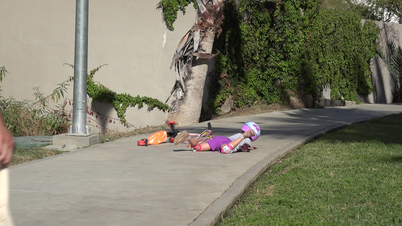 The fallen child remains beside the orange scooter while part of an adult enters the left edge of the San Jose Del Cabo sidewalk scene.