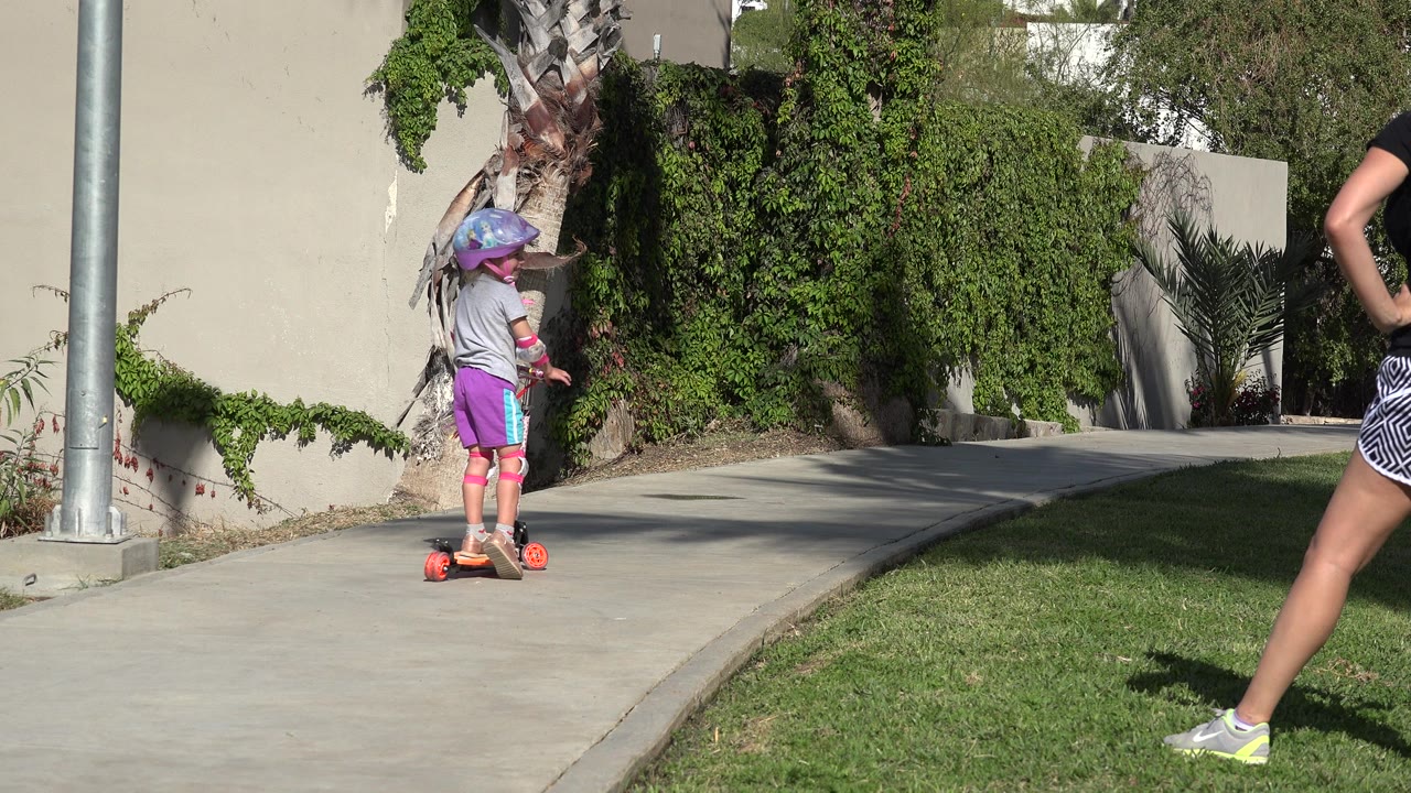 Young child on a scooter beside grass and a stucco wall.