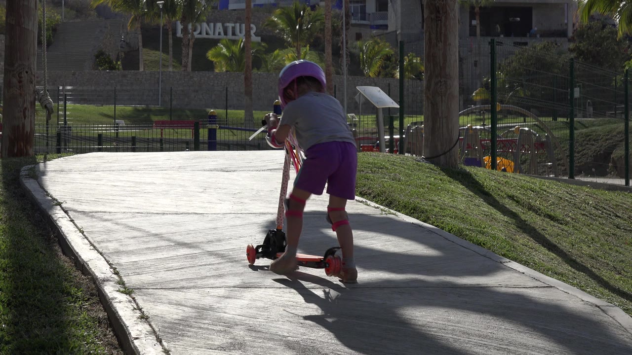 A young girl in a purple helmet and knee pads begins riding a red scooter away from camera on a sunlit path in San Jose Del Cabo, Mexico.