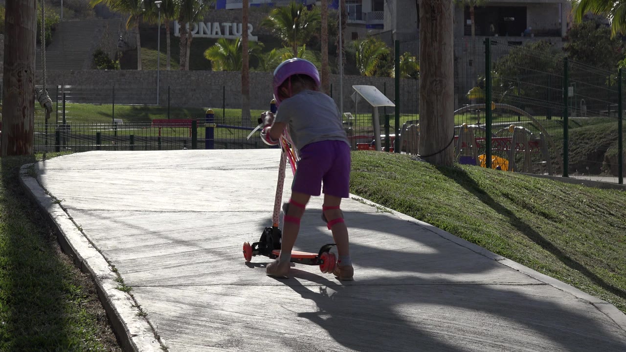 The child balances on the red scooter near the center of the paved path, with strong sunlight reflecting off the walkway and palm trees in the background.