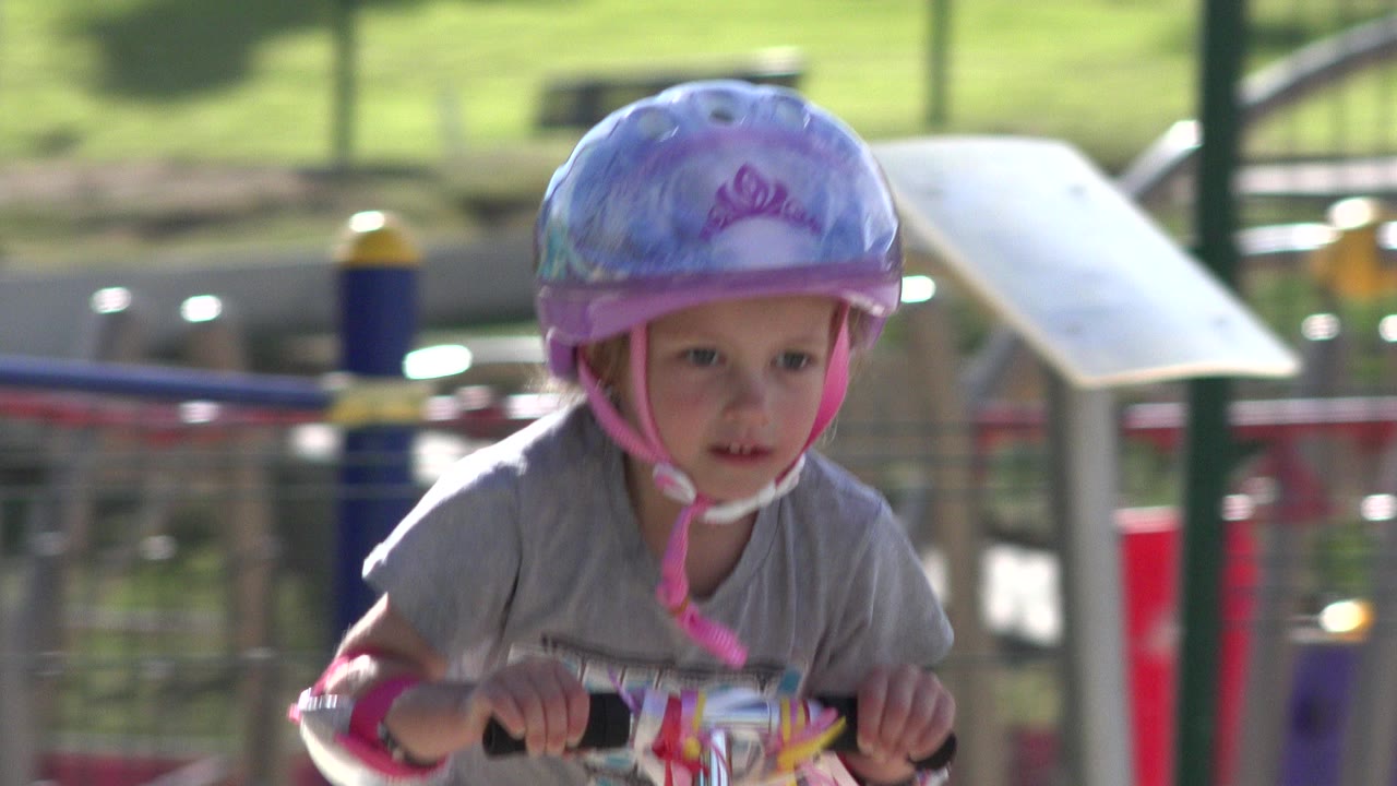 Little girl in a purple helmet riding a scooter near playground equipment.