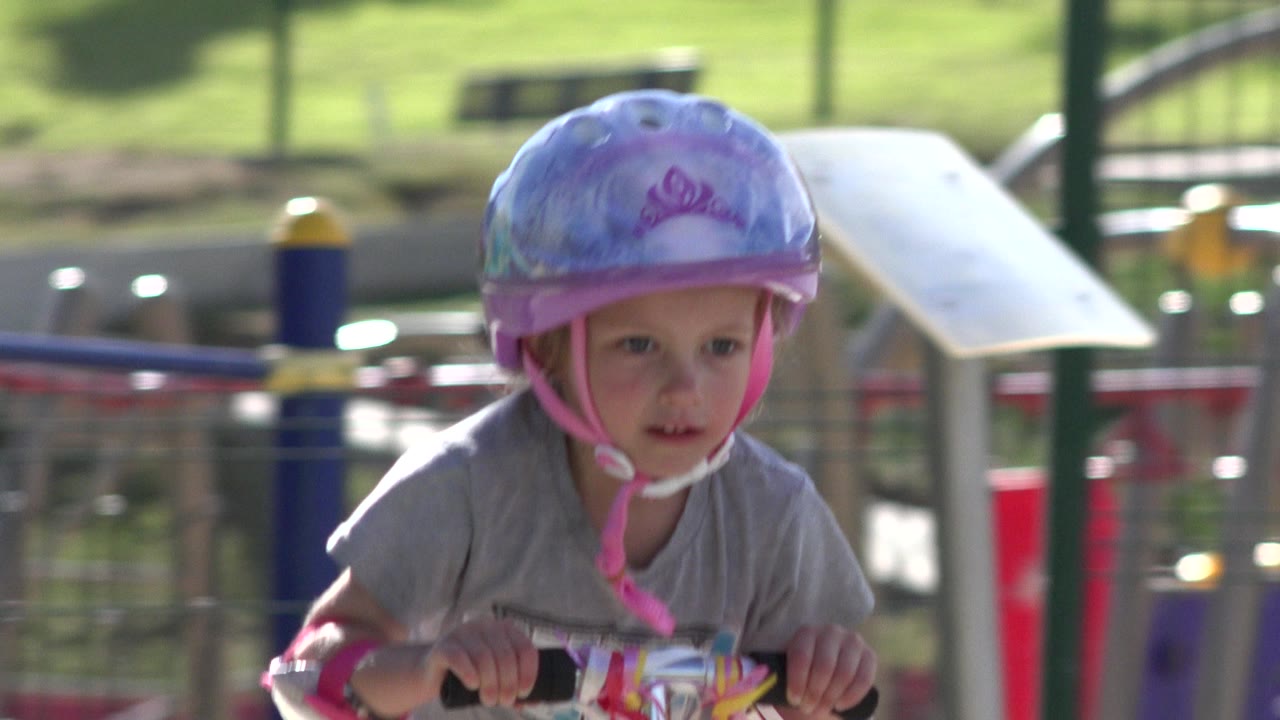 A little girl in a purple helmet rides her scooter toward the camera in San Jose del Cabo, with soft playground shapes and green lawn in the background.