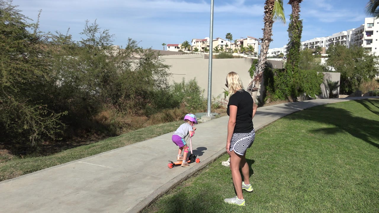 Child on an orange scooter with a woman watching beside a sunny path.