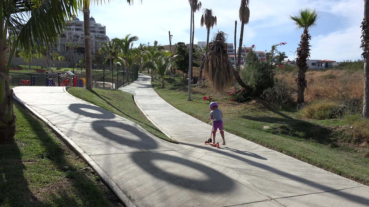 The child continues on the scooter in San Jose Del Cabo, Mexico, positioned near the right side of the path as palm shadows cross the concrete and grass borders both sides.