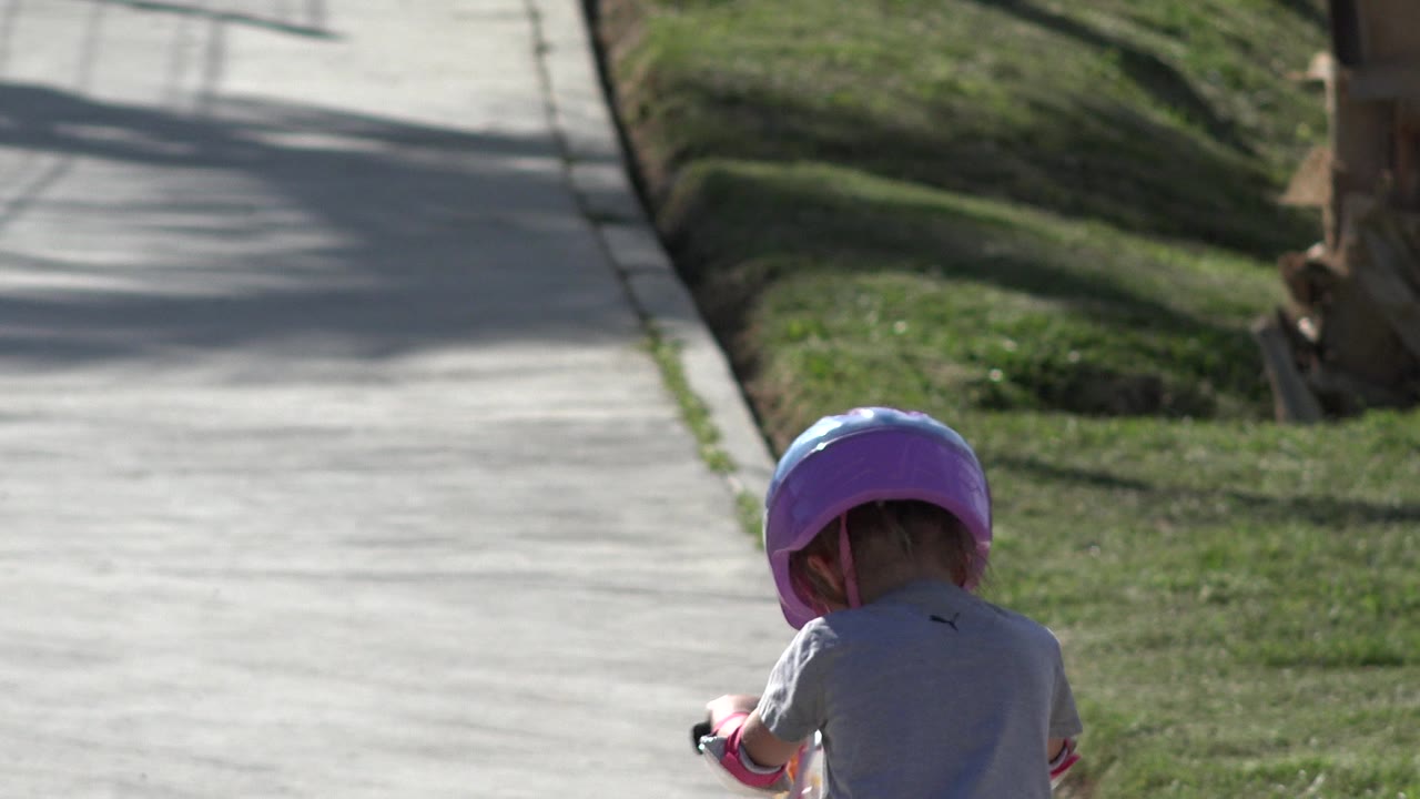 Child in a purple helmet walking a scooter on a paved park path.