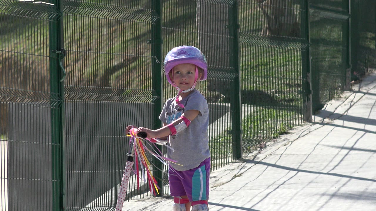 Child in helmet holding a scooter beside a tall fence.