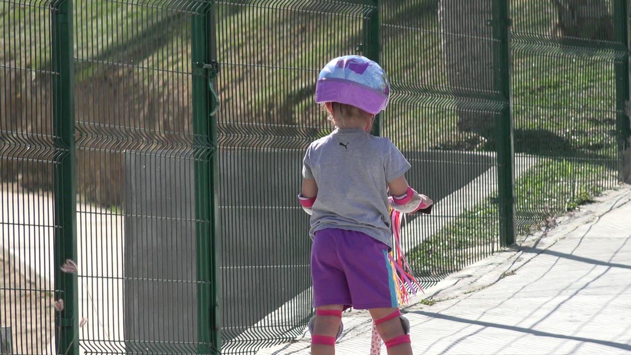 Child in purple shorts and helmet rides a scooter toward a green gate.