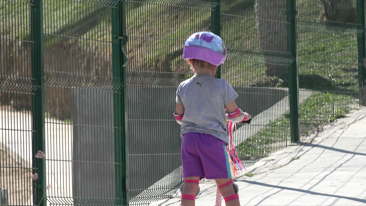 At 0:01, the child remains centered from behind while the scooter and green gate frame a sunlit paved path in San Jose del Cabo, Mexico.